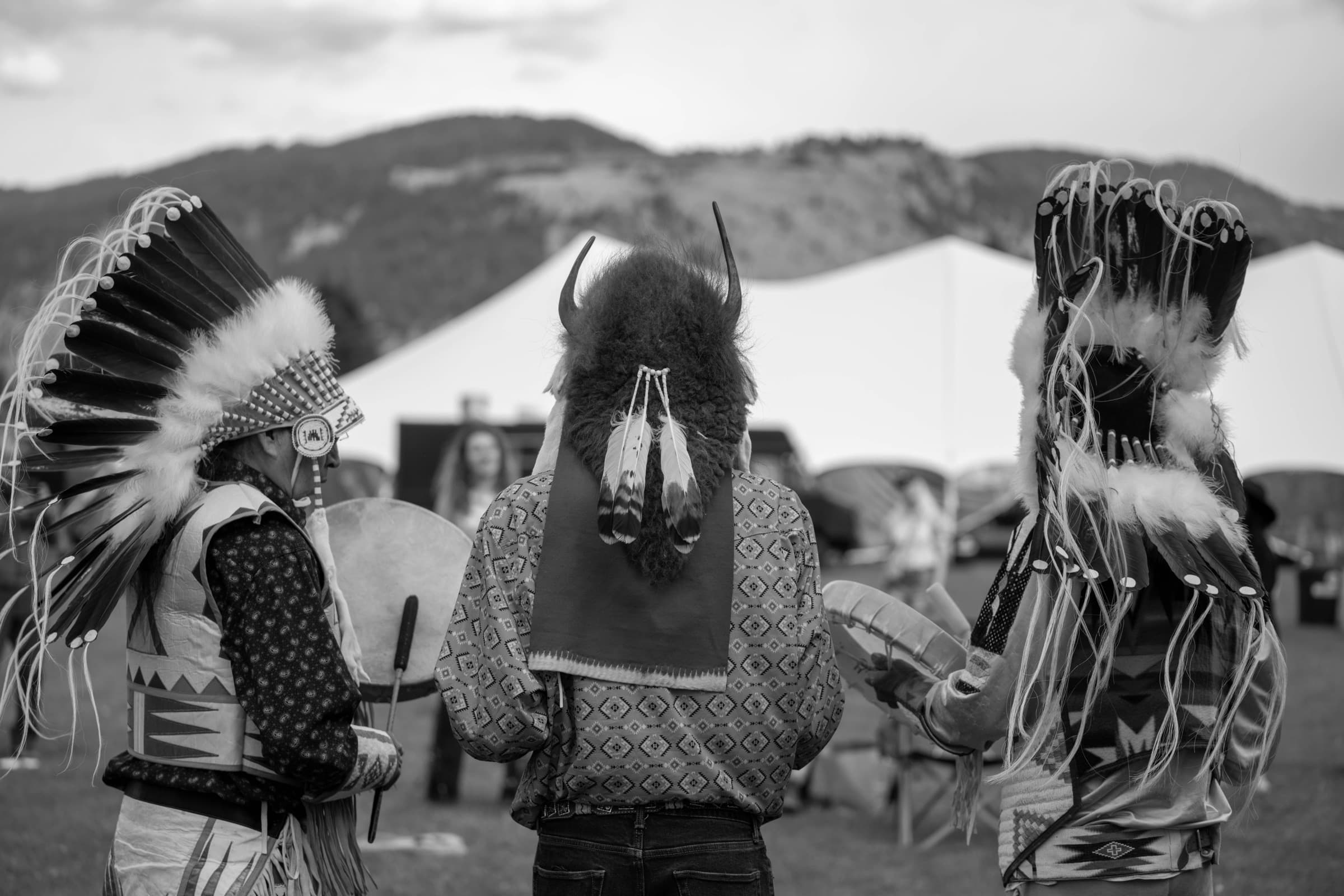 Three dancers in full regalia at the Horseshoe Music Festival, seen from behind, with the Tetons and the festival tents beyond