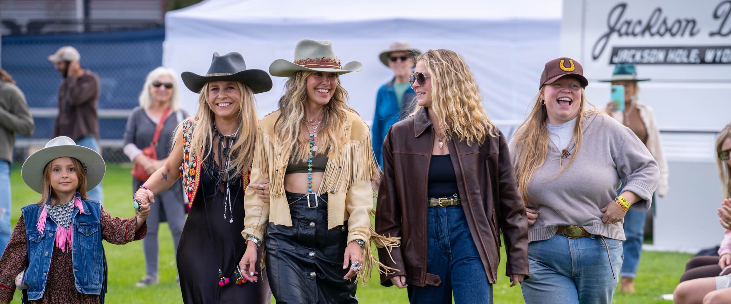 Four women and a small girl walking arm in arm across the grass at Horseshoe, laughing, in hats and boots with the tents behind them