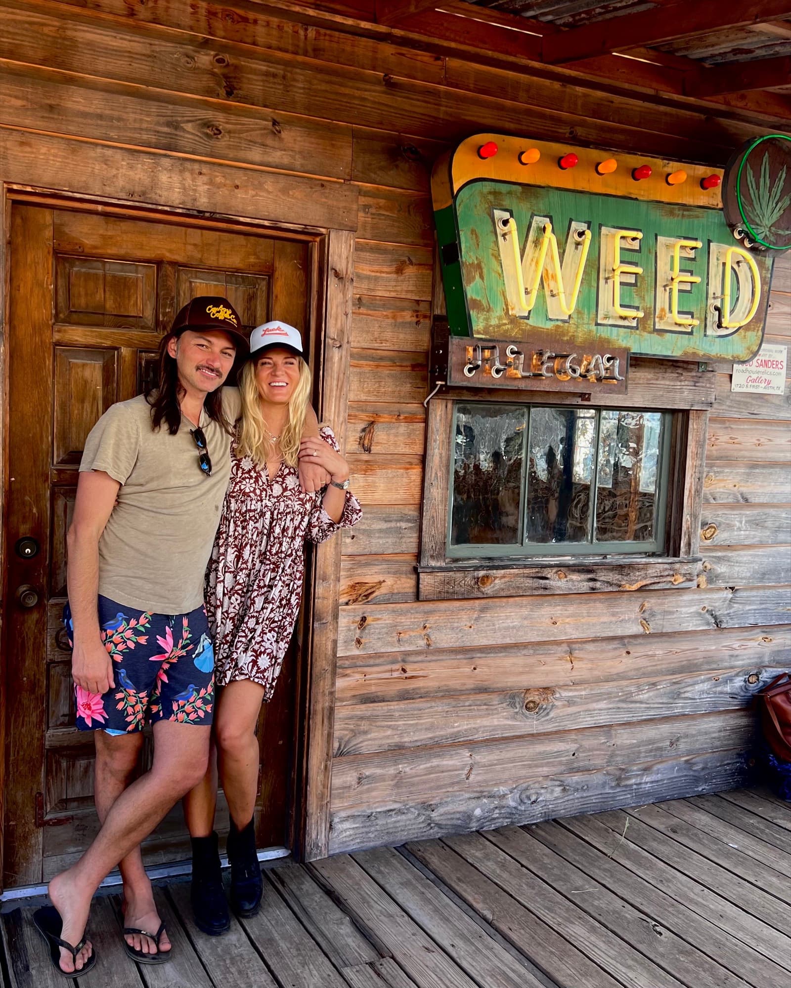 Duane and Lisa Betts on the porch of a weathered timber building, arms around each other and both smiling broadly, in ball caps on a bright day; a large vintage neon sign reading WEED, with a cannabis leaf mounted beside it, hangs on the wall to their right