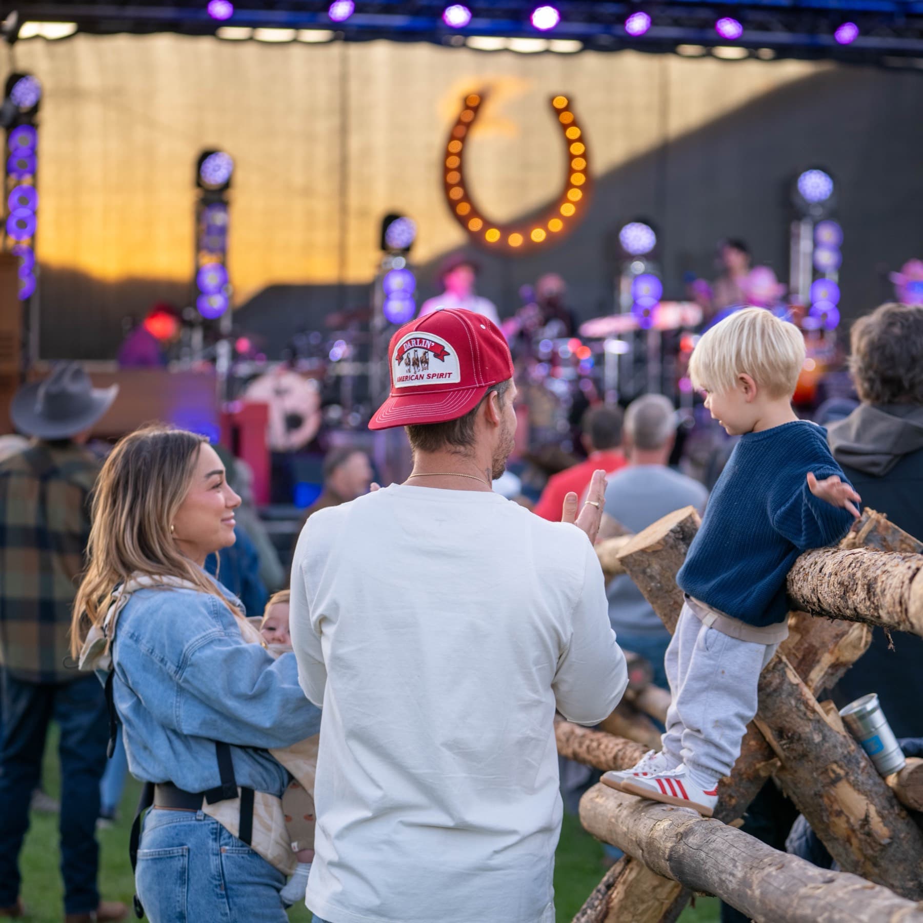 A couple with a baby in a carrier and a small boy standing up on the log fence beside them, all watching the band under the lit horseshoe at dusk
