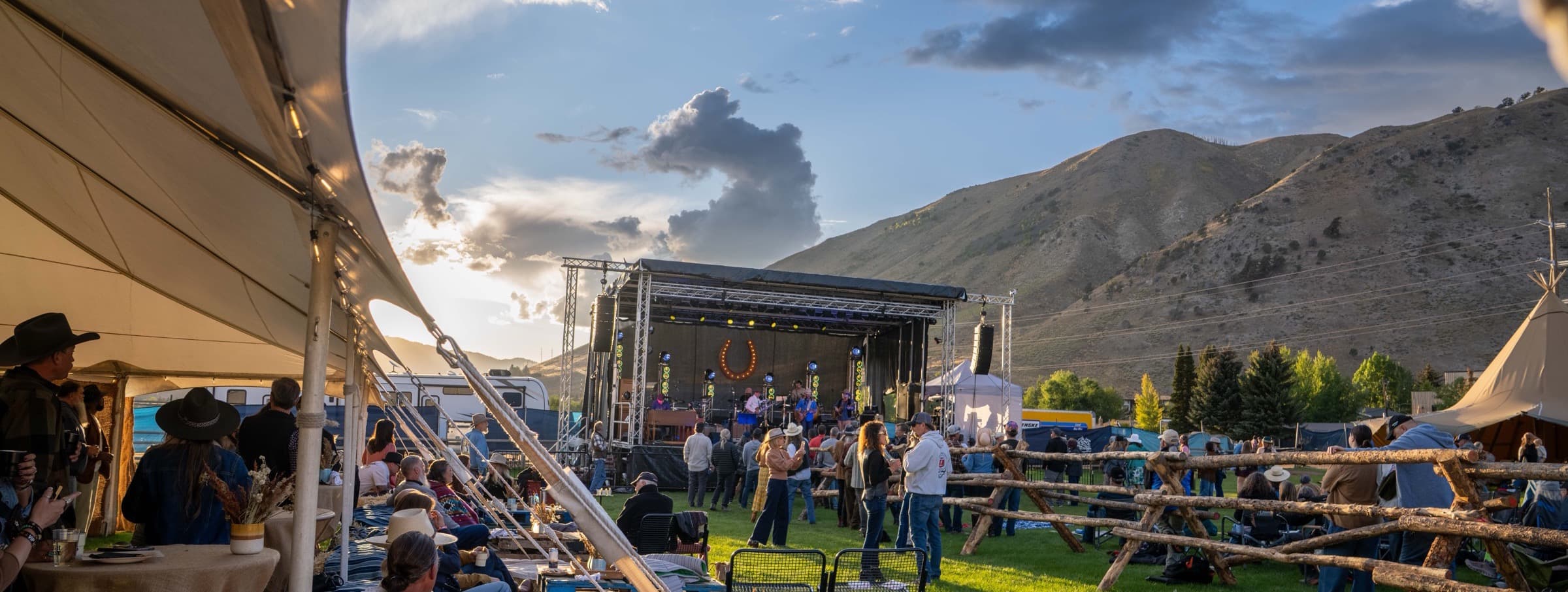 The Horseshoe grounds at golden hour, a canvas tipi and hay-bale seating in the foreground and the festival stage against the mountains beyond