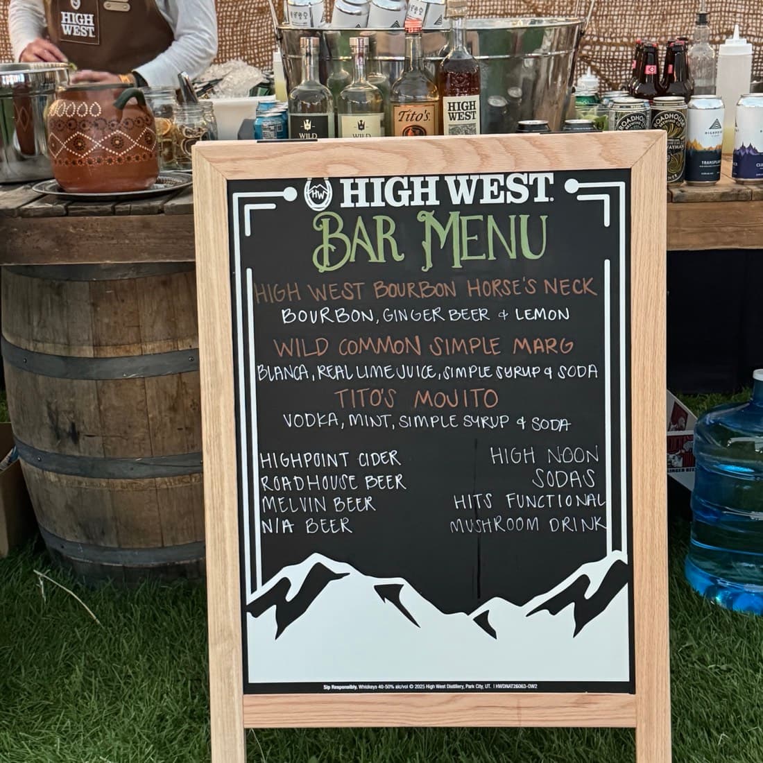 A chalkboard bar menu propped on a barrel, bottles and glassware behind it