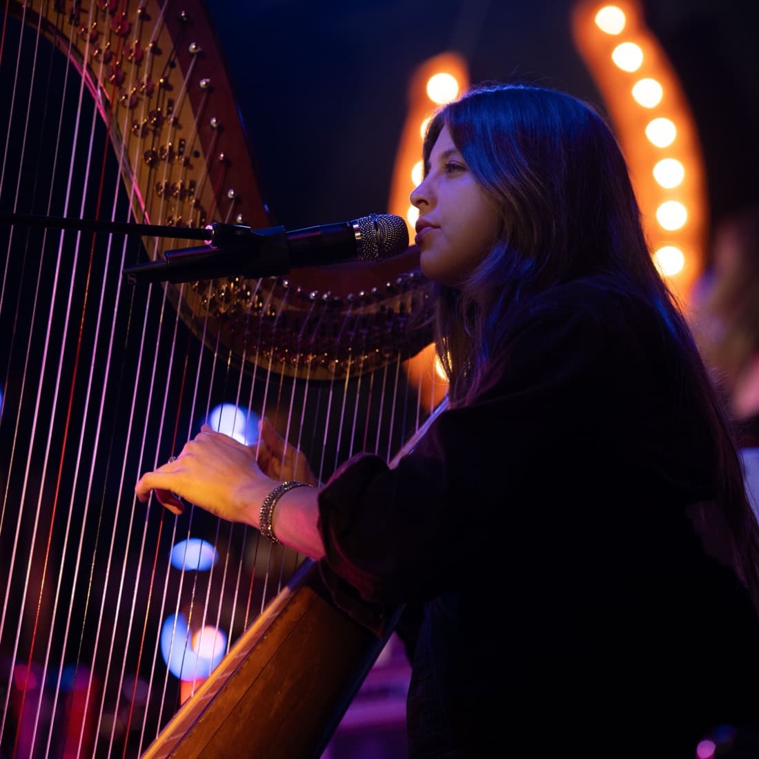 A harpist singing into a microphone as she plays, the strings lit from behind