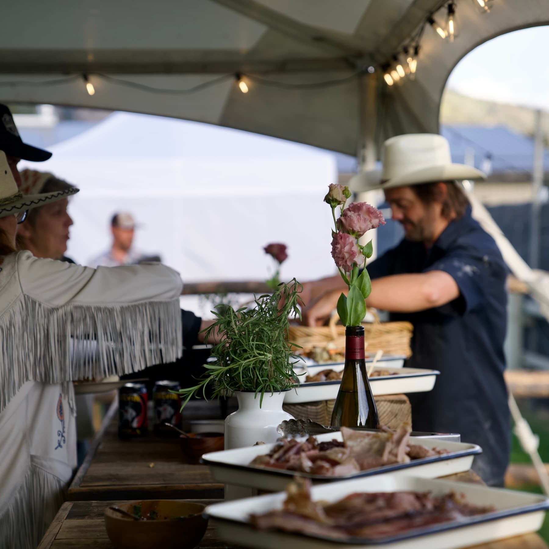 Cooks plating at a long service table under an open-sided tent, flowers and herbs between the dishes