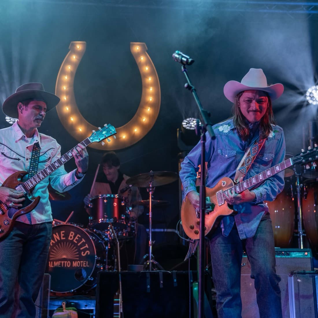 Two guitarists in cowboy hats playing side by side in front of the lit horseshoe backdrop