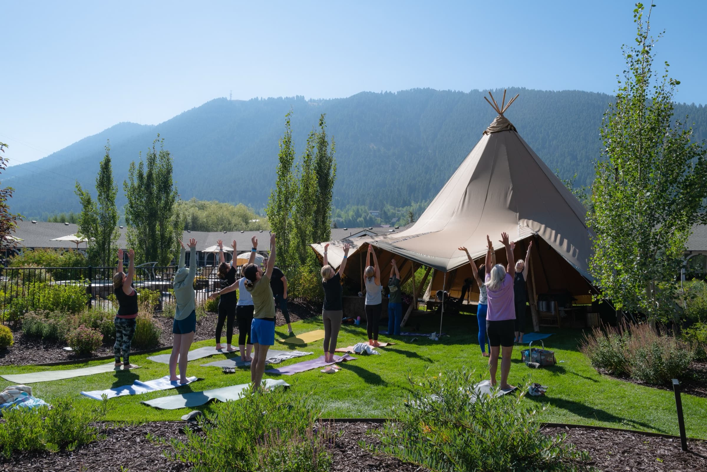 A morning class on the grass in front of the tipi at Horseshoe, a dozen people on mats with their arms raised and a forested mountain behind