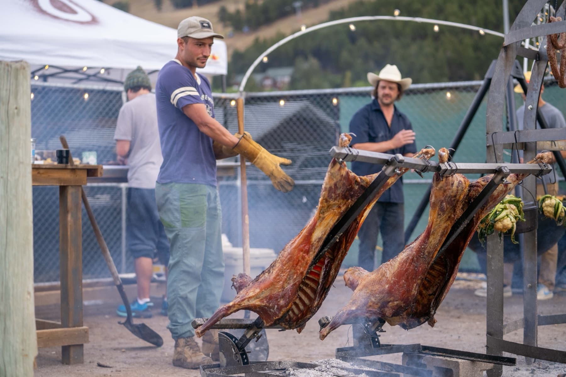 Two cooks tending whole cuts of meat roasting on iron crosses over an open fire