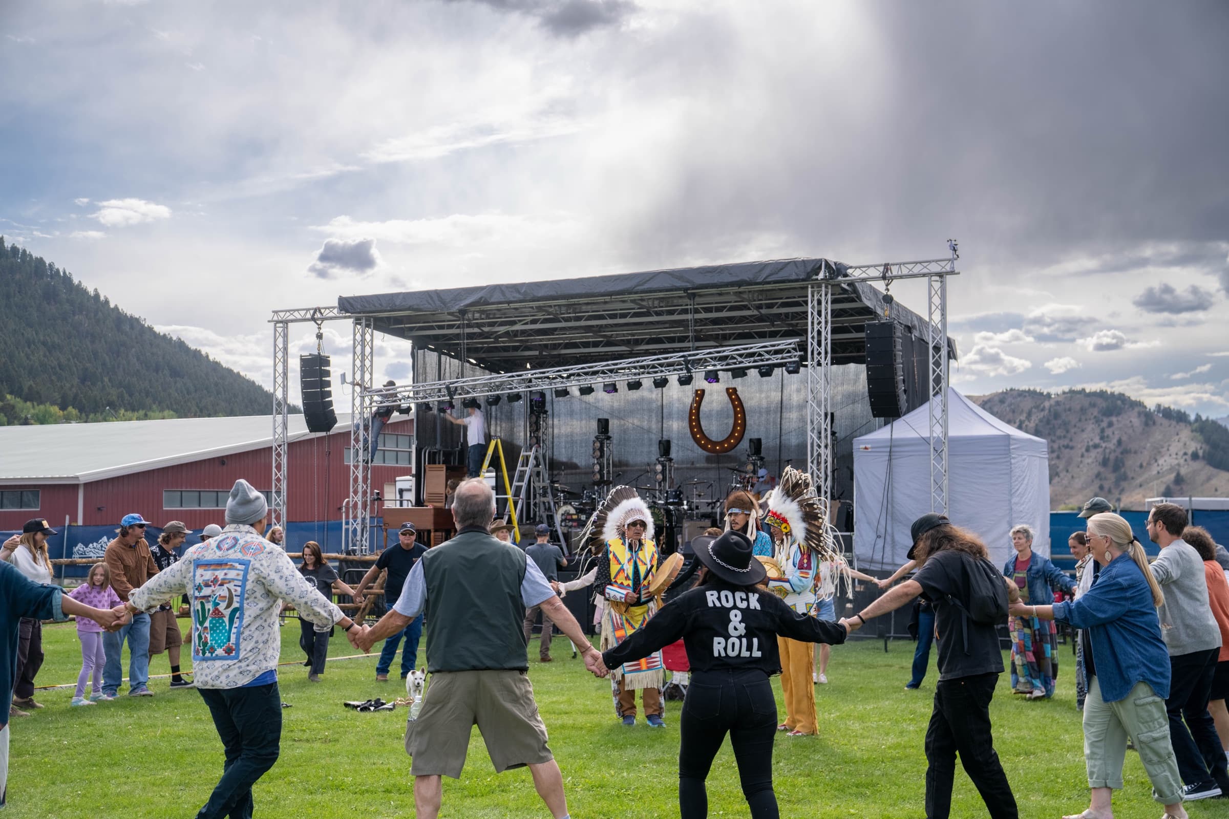 A circle of people holding hands on the grass in front of the Horseshoe stage, dancers in regalia among them