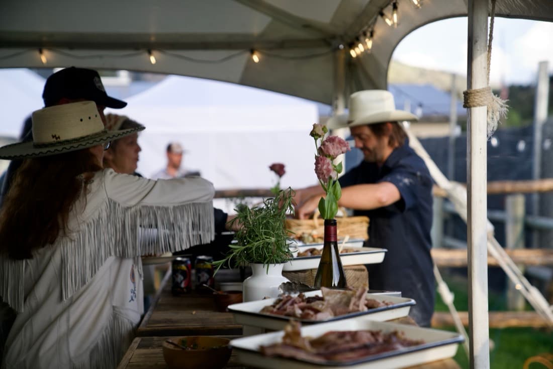 Two people serving from platters on a flower-dressed table under a tent