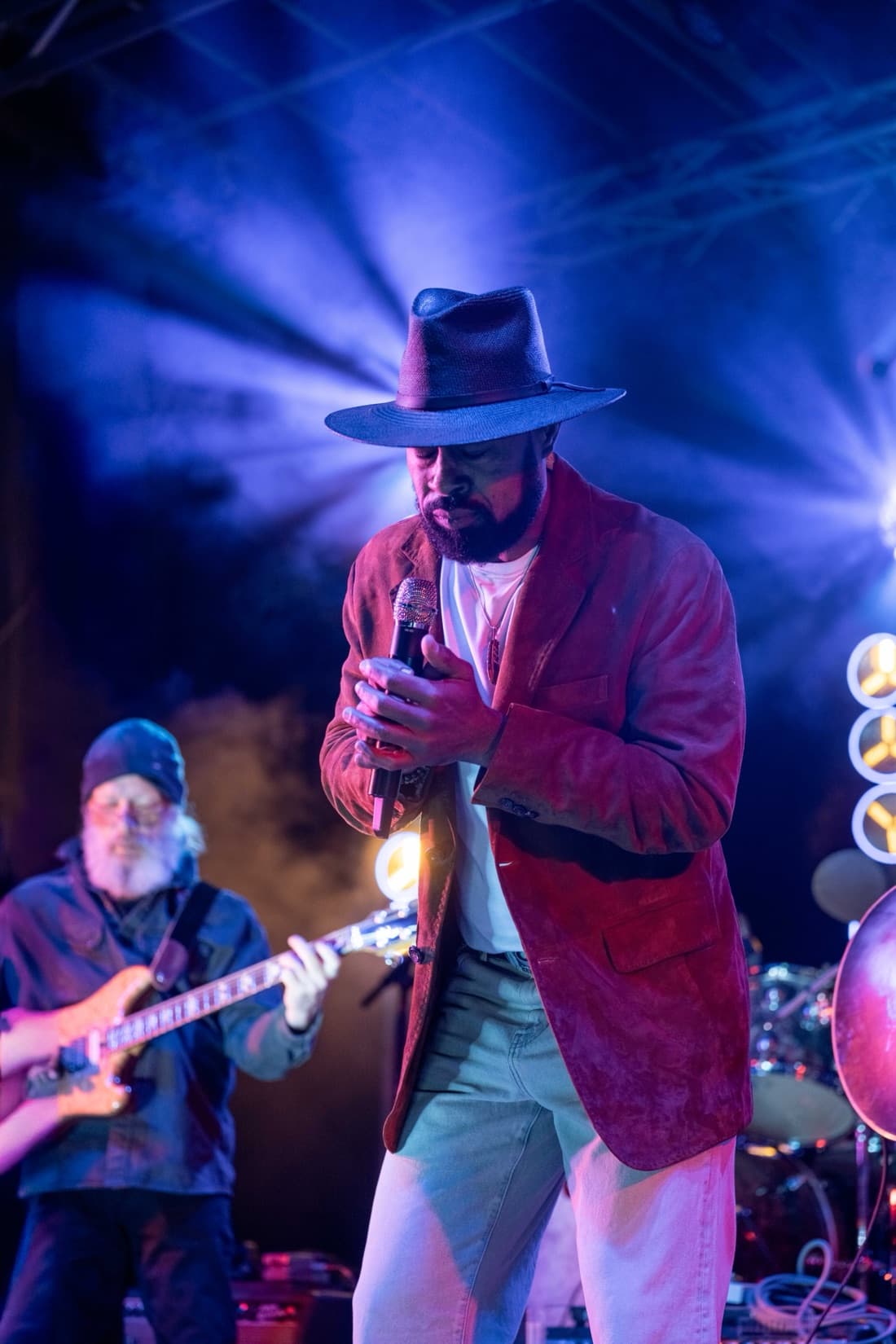 A singer in a wide-brimmed hat and red jacket at the microphone under blue stage light, a guitarist behind him