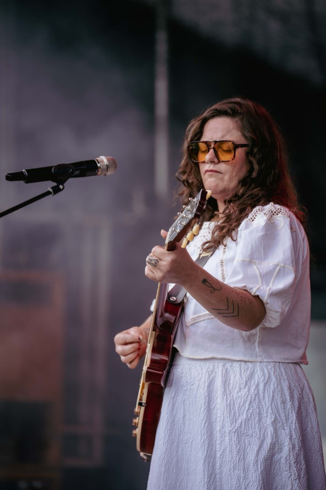 A musician in a white dress and sunglasses singing at the microphone with an electric guitar