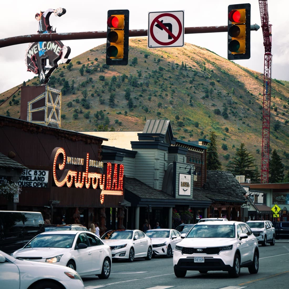 The Million Dollar Cowboy Bar sign lit on Jackson town square at dusk, traffic queued beneath the neon welcome rider and Snow King rising behind